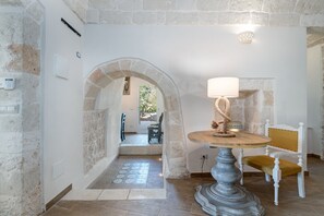 Una habitación con pared de piedra, una mesa redonda con una lámpara, una silla blanca y un arco que da a otra estancia.