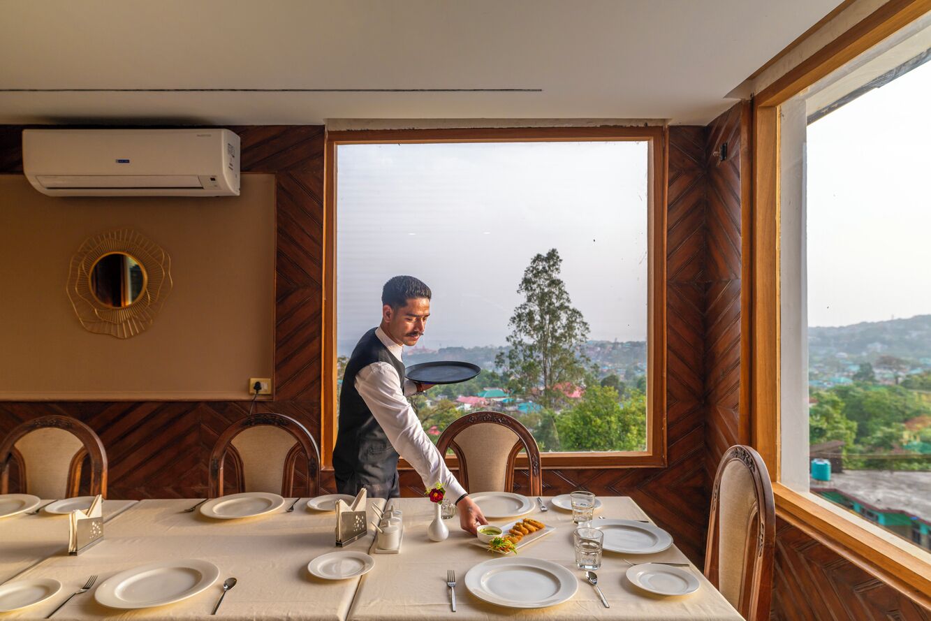 Een eetzaal met een gedekte tafel, een ober die eten serveert, en een groot raam met uitzicht op groen.