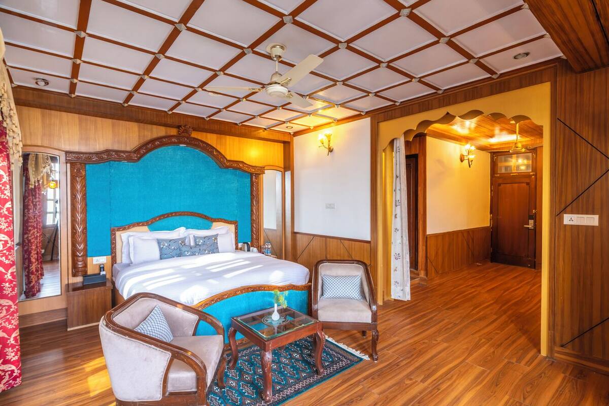 Quarto de hotel com uma cama grande, duas cadeiras, uma mesinha, ventoinha no teto e piso de madeira.