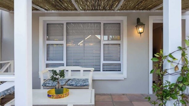 Un patio cubierto con una mesa y sillas blancas, una ventana con persianas y un aplique de pared.