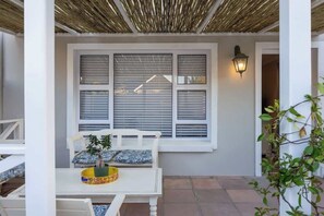 Ein überdachter Patio-Bereich mit einem weißen Tisch und Stühlen, einem Fenster mit Jalousien und einer Wandleuchte.