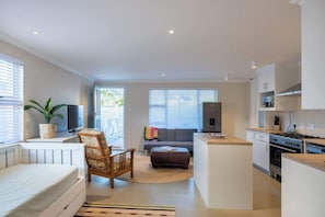 Une cuisine moderne avec des meubles blancs, un coin repas comprenant une table et des chaises, un canapé et une grande fenêtre avec des stores.