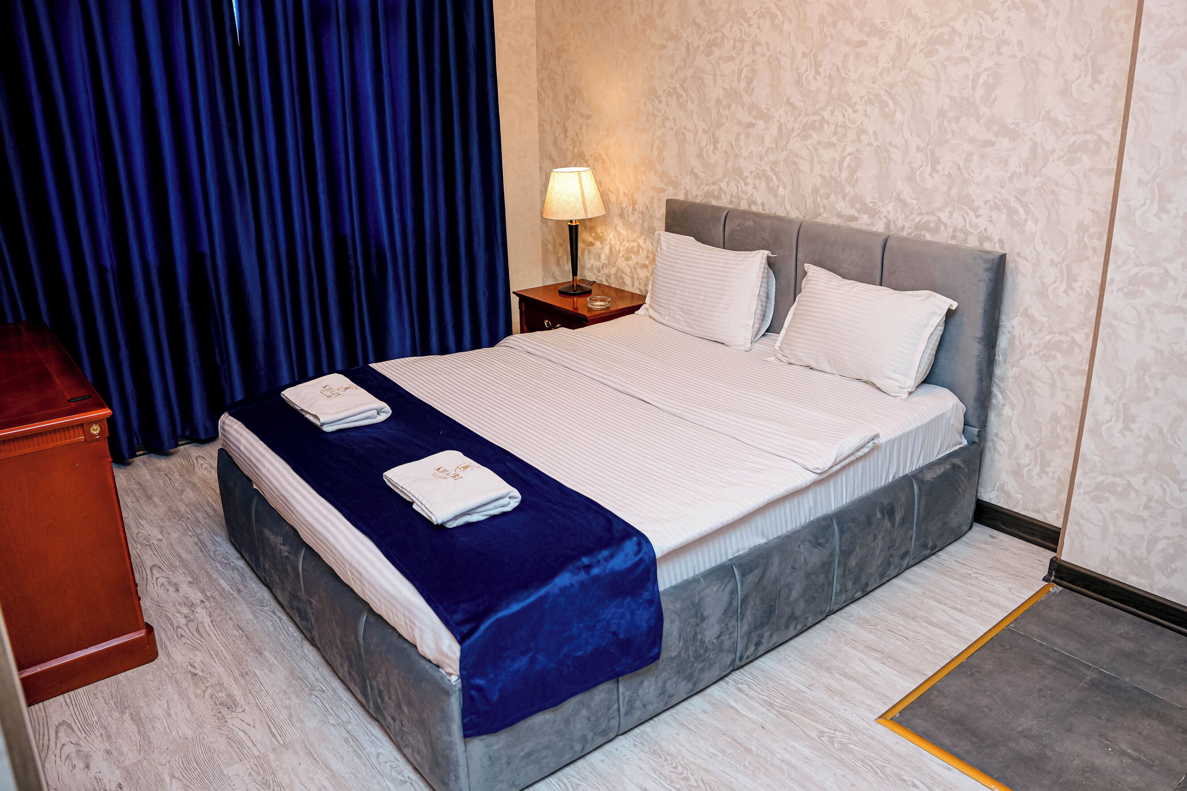 Una habitación de hotel con cama, mesita de noche, lámpara y cortina azul.