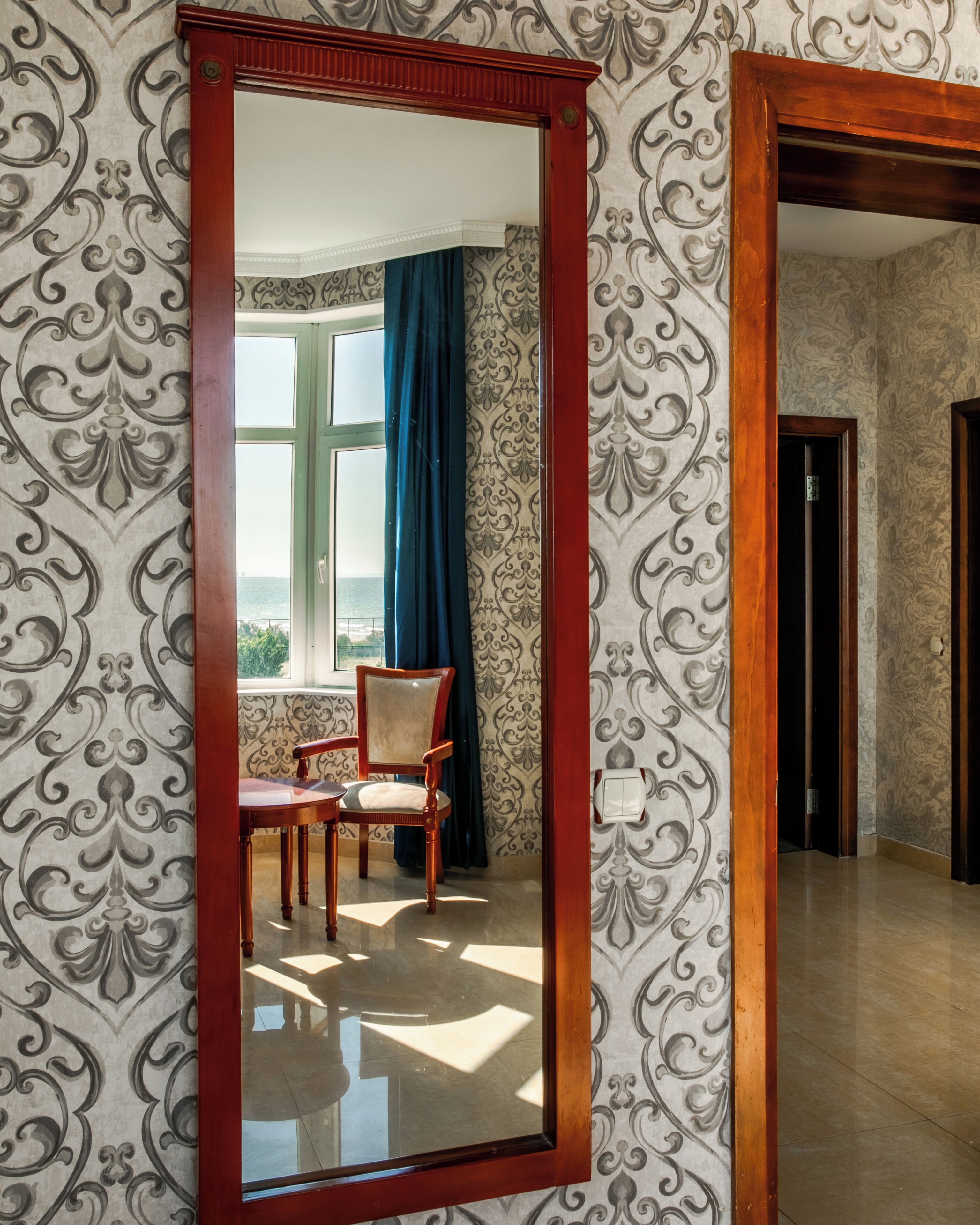 Ein Zimmer mit einem großen Spiegel, strukturiertem Wandpapier, einem Stuhl und Blick ins Freie durch ein Fenster.