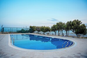 Una piscina con solárium de baldosas, sillas de descanso y sombrillas, rodeada de árboles y con vistas a una masa de agua.