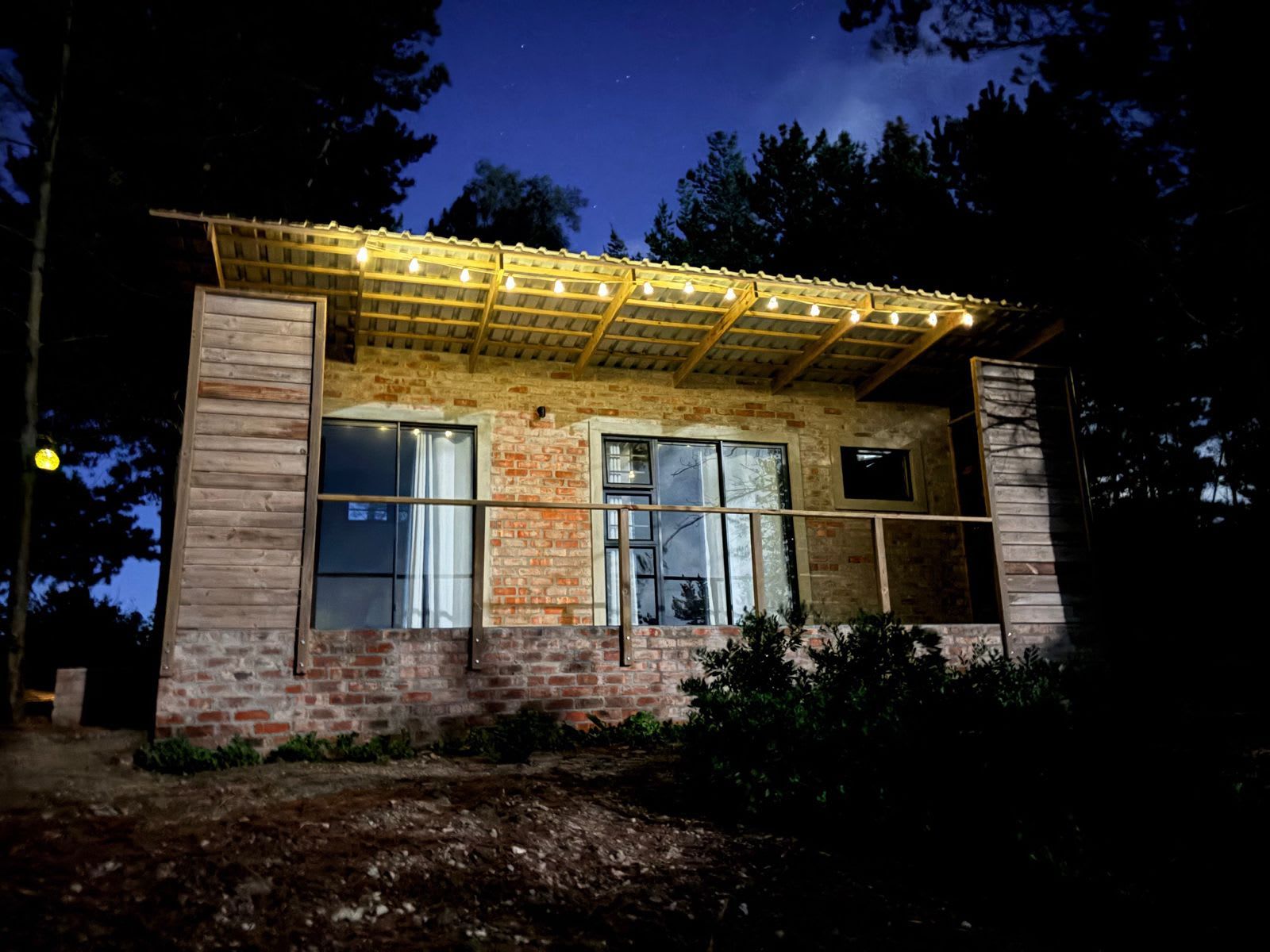 Un edificio rústico con fachada de ladrillo, amplias ventanas y una pérgola de madera con luces.
