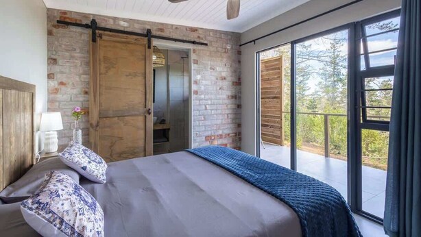 A bedroom with a bed, a ceiling fan, a large window, and a brick wall.
