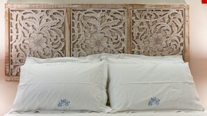 Cama com lençóis brancos e duas almofadas com texto bordado, diante de uma parede com cabeceira de madeira entalhada.
