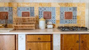 Uma cozinha com painel de azulejos, armários de madeira, fogão a gás, um varal de madeira e uma panela de cerâmica.