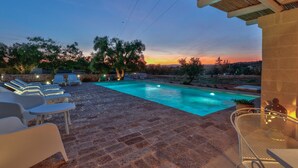 Piscina com espreguiçadeiras e pérgula, cercada por árvores e vista para o pôr do sol.