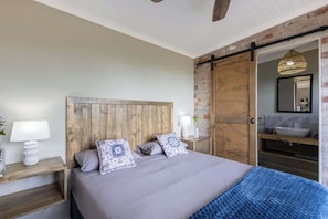 A bedroom with a wooden headboard, a large bed, a nightstand with a lamp, a bathroom with a sink and mirror, and a hanging light fixture.
