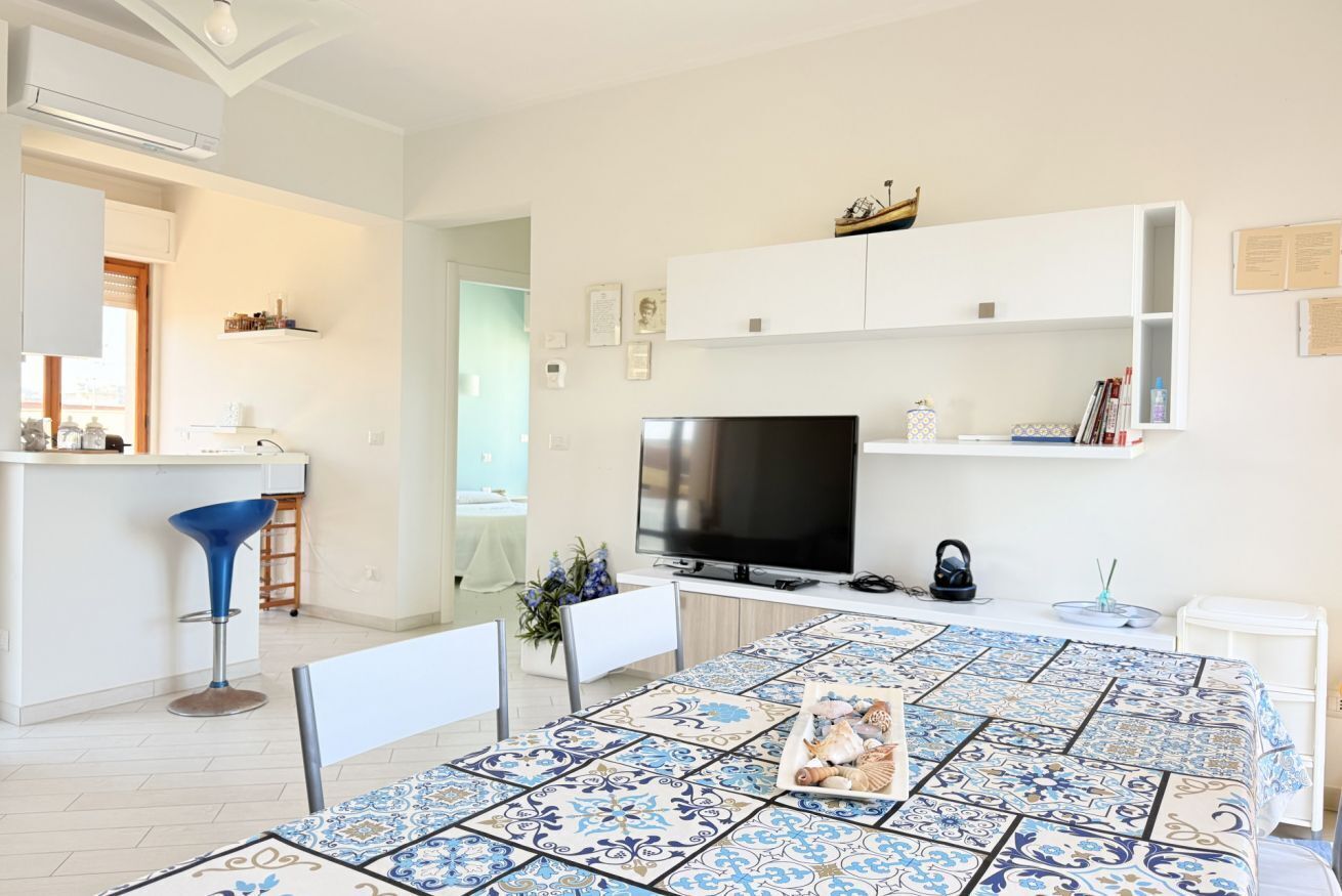 Una sala de estar moderna con un televisor, estanterías blancas, una mesa de comedor con mantel estampado y una zona de bar con una silla azul.