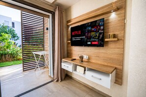 Ein modernes Wohnzimmer mit einem an der Wand montierten Fernseher, der Netflix zeigt, einem Holz-TV-Möbel, einem Glastisch und einer Schiebetür zu einem Außenbereich.