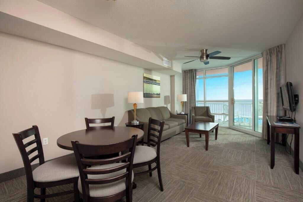 A hotel room with a dining table, chairs, a sofa, a TV, and a balcony with a view.