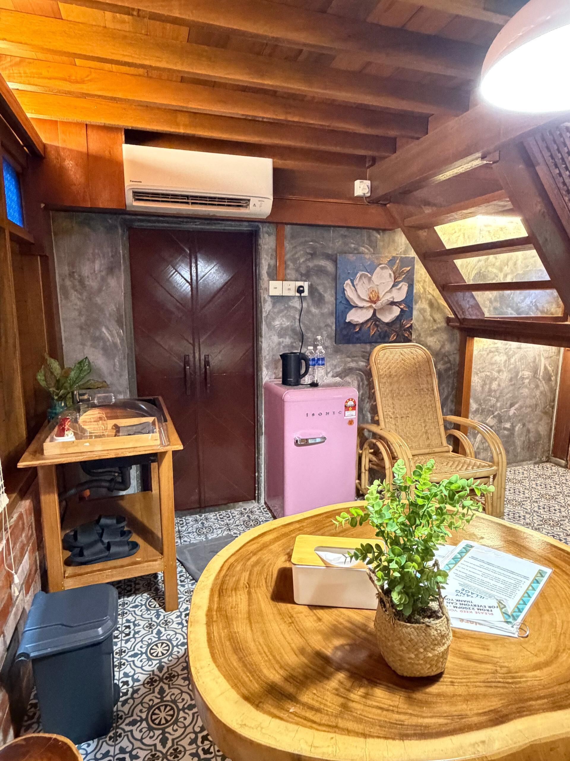 A cozy room with a wooden ceiling, a large window, a wooden table with a plant, a chair, and a small pink refrigerator.