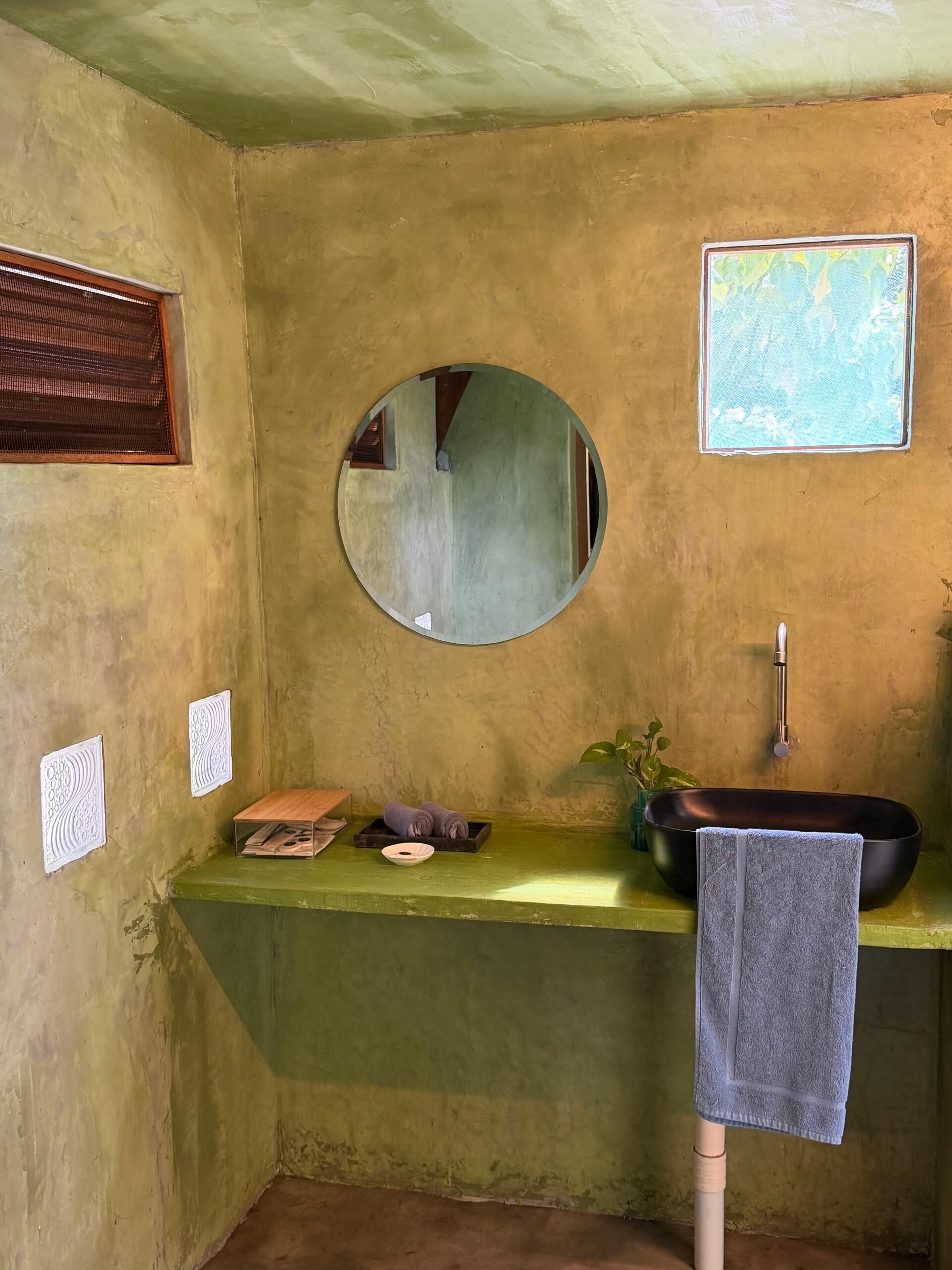 Ein Badezimmer mit rundem Spiegel, einem Waschbecken, einem Handtuchhalter und einem kleinen Regal mit Handtüchern und einer Schüssel.