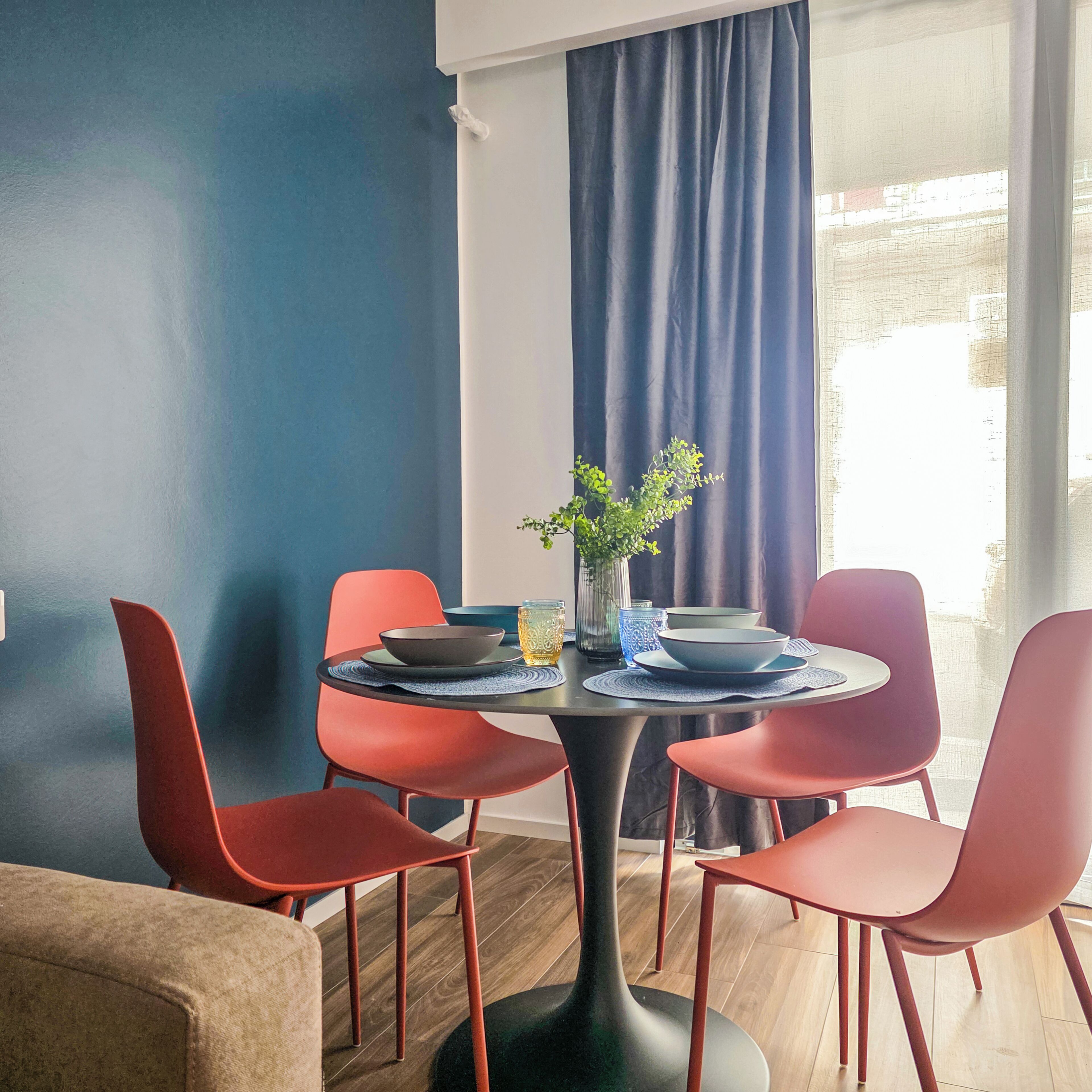 A dining area with a round table, red chairs, a vase with a plant, and a view of the outdoors through a window with blue curtains.
