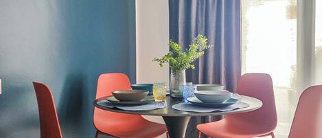 A dining area with a round table, red chairs, a vase with a plant, and a view of the outdoors through a window with blue curtains.