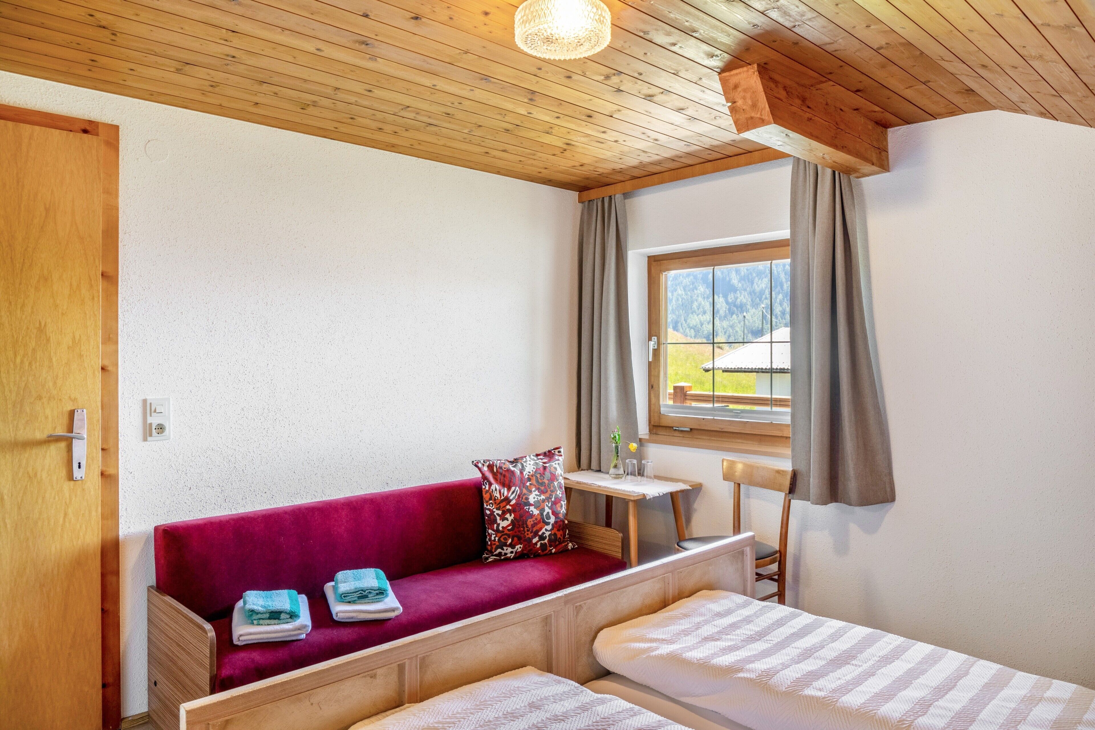 Una habitación con techo de madera, un sofá rojo con un cojín estampado, una mesita con un florero y una ventana con cortinas.