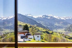 Une maison moderne avec une voiture garée devant, entourée de collines verdoyantes et, au loin, de montagnes aux sommets enneigés.