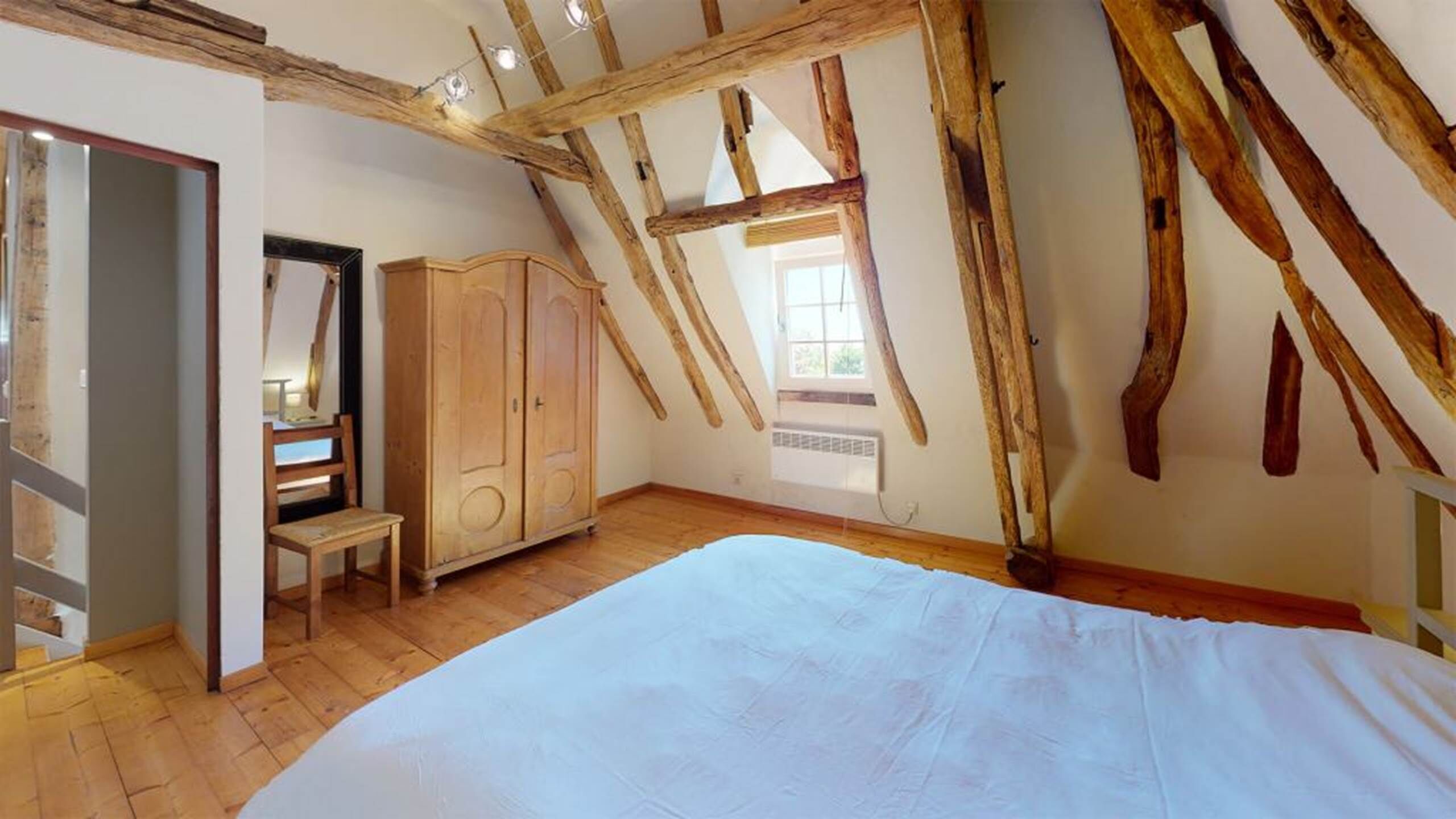 Un dormitorio con techo inclinado, vigas de madera, una cama, un armario y una ventana.