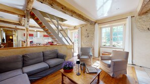 A modern living room with a wooden staircase, a sofa, two armchairs, a table with a plant, and a view of the kitchen and garden through a large window.