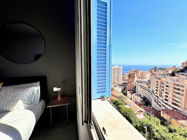 Ein Hotelzimmer mit einem Bett, einem kleinen Tisch, einem Nachttisch und einem Fenster mit Blick auf die Stadt und das Meer.
