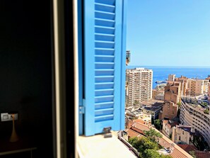 Blick durch ein Fenster mit blauen Fensterläden auf eine Küstenstadt mit Gebäuden und dem Meer.