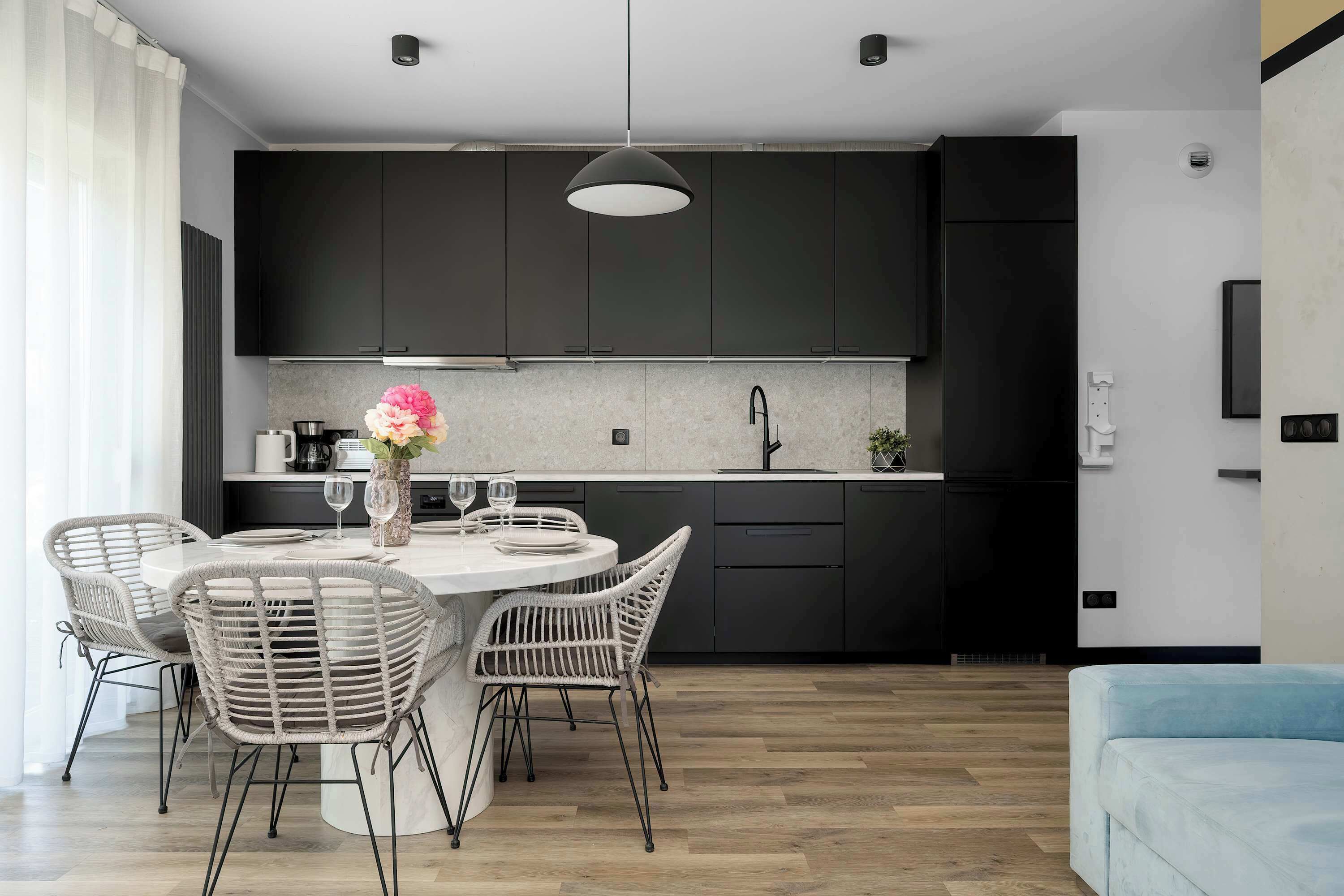 Een moderne keuken met donkere kastjes, een ronde eettafel met rieten stoelen, en een lichtblauwe bank in de woonkamer.