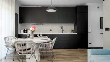 Een moderne keuken met donkere kastjes, een ronde eettafel met rieten stoelen, en een lichtblauwe bank in de woonkamer.