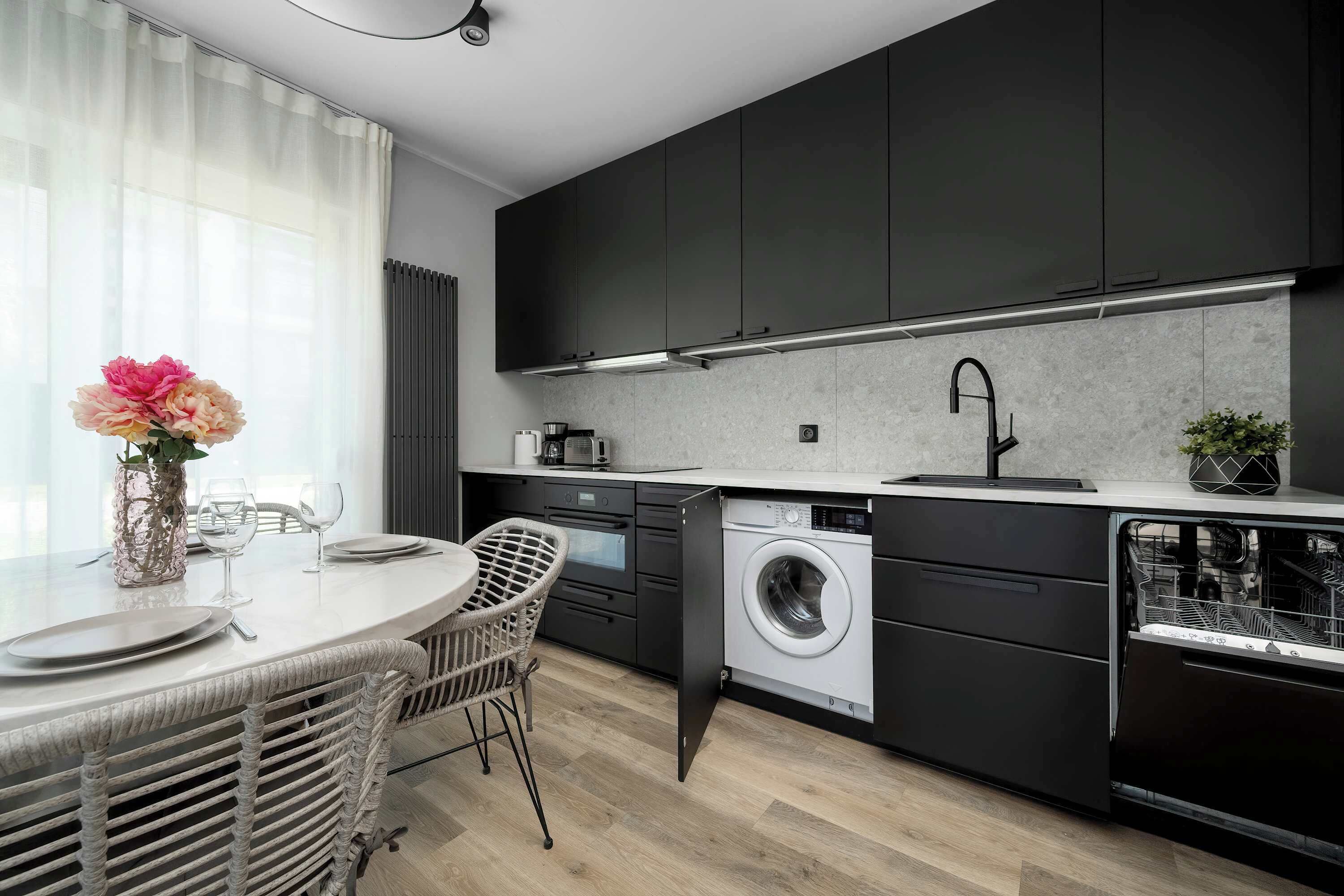 Una cocina moderna con armarios oscuros, lavadora integrada y una zona de comedor con sillas de mimbre.
