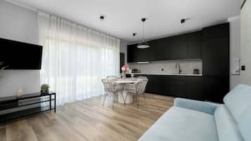 Een moderne keuken met donkere kastjes, een eethoek met een ronde tafel en stoelen, een lichtblauwe bank en een flatscreen televisie die aan de muur is bevestigd.