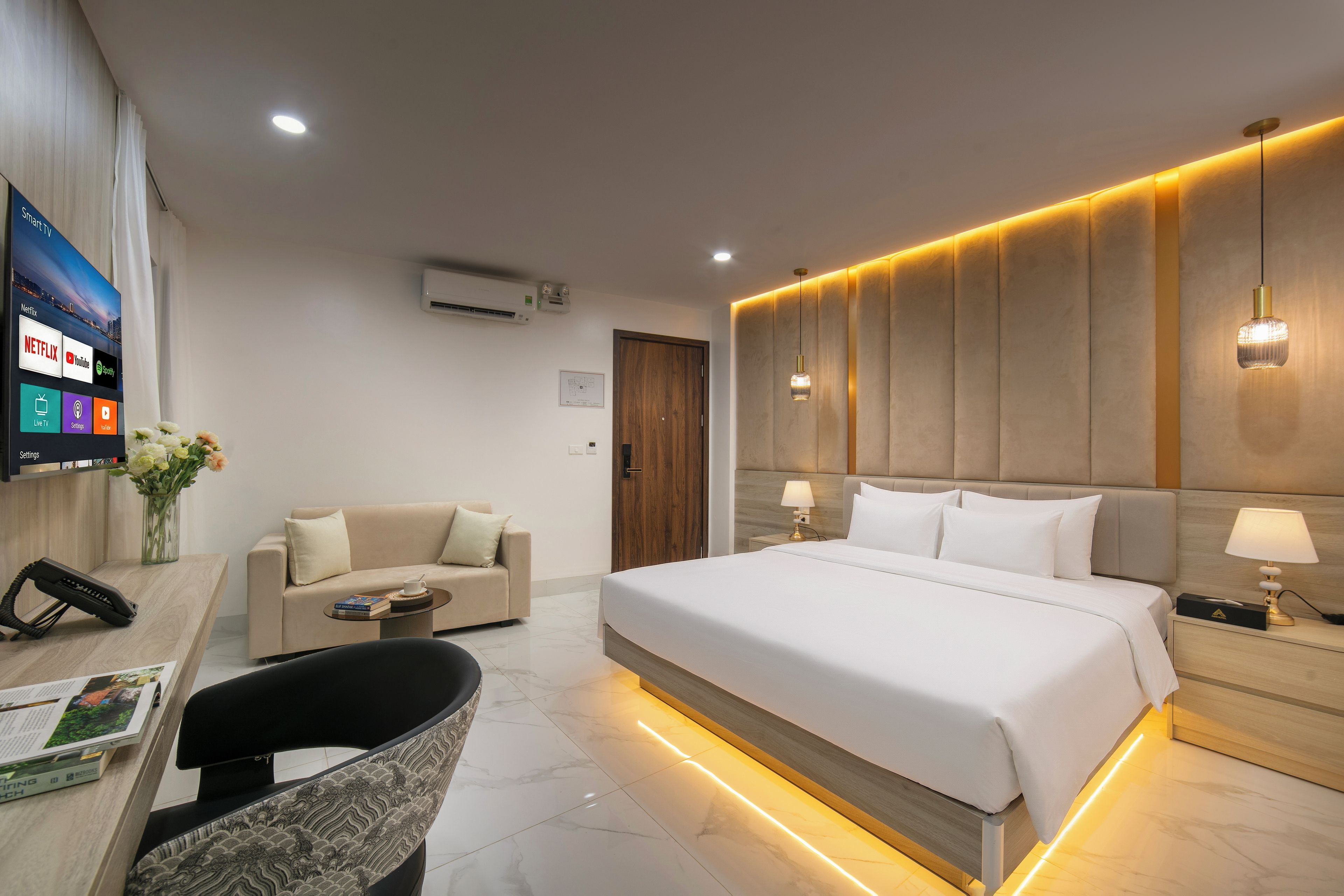 A hotel room with a bed, a sofa, a desk with a phone, a TV displaying streaming services, and a wall-mounted air conditioner.