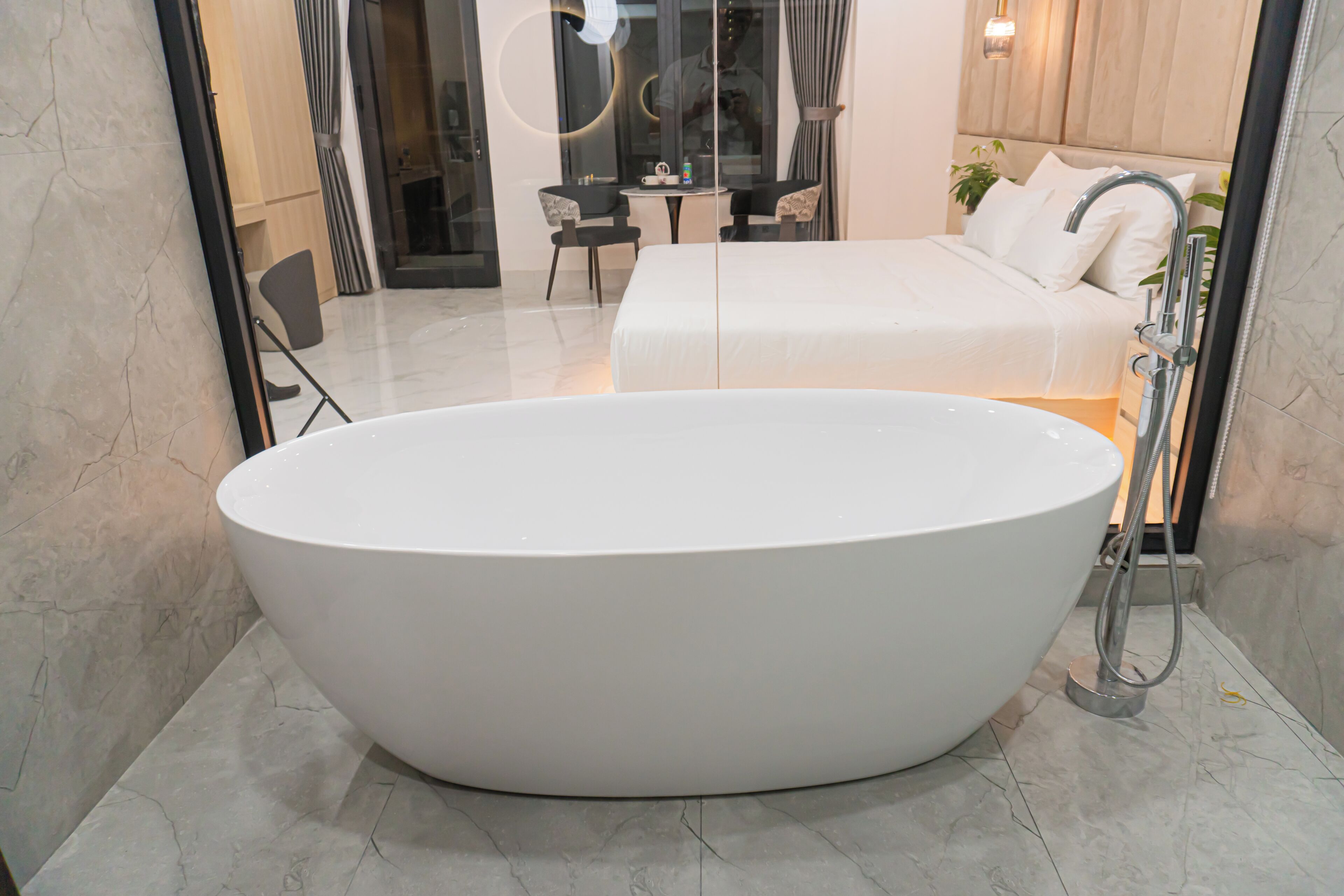 A modern bathroom with a large oval bathtub, a separate shower area, and a visible bedroom with a bed and a nightstand.