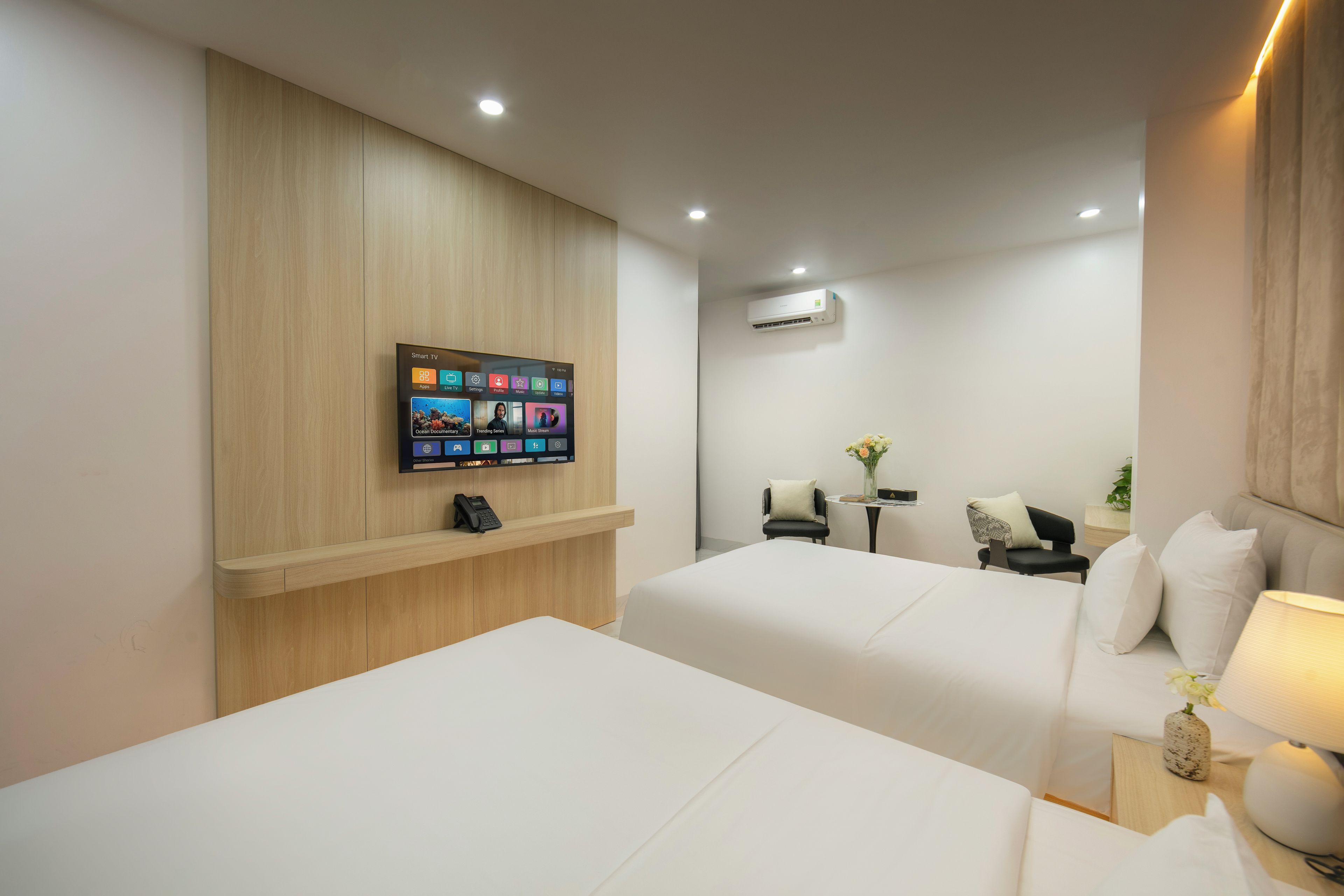 Habitación de hotel con dos camas, un televisor montado que muestra varios canales, un escritorio con una lámpara y un equipo de aire acondicionado en la pared.