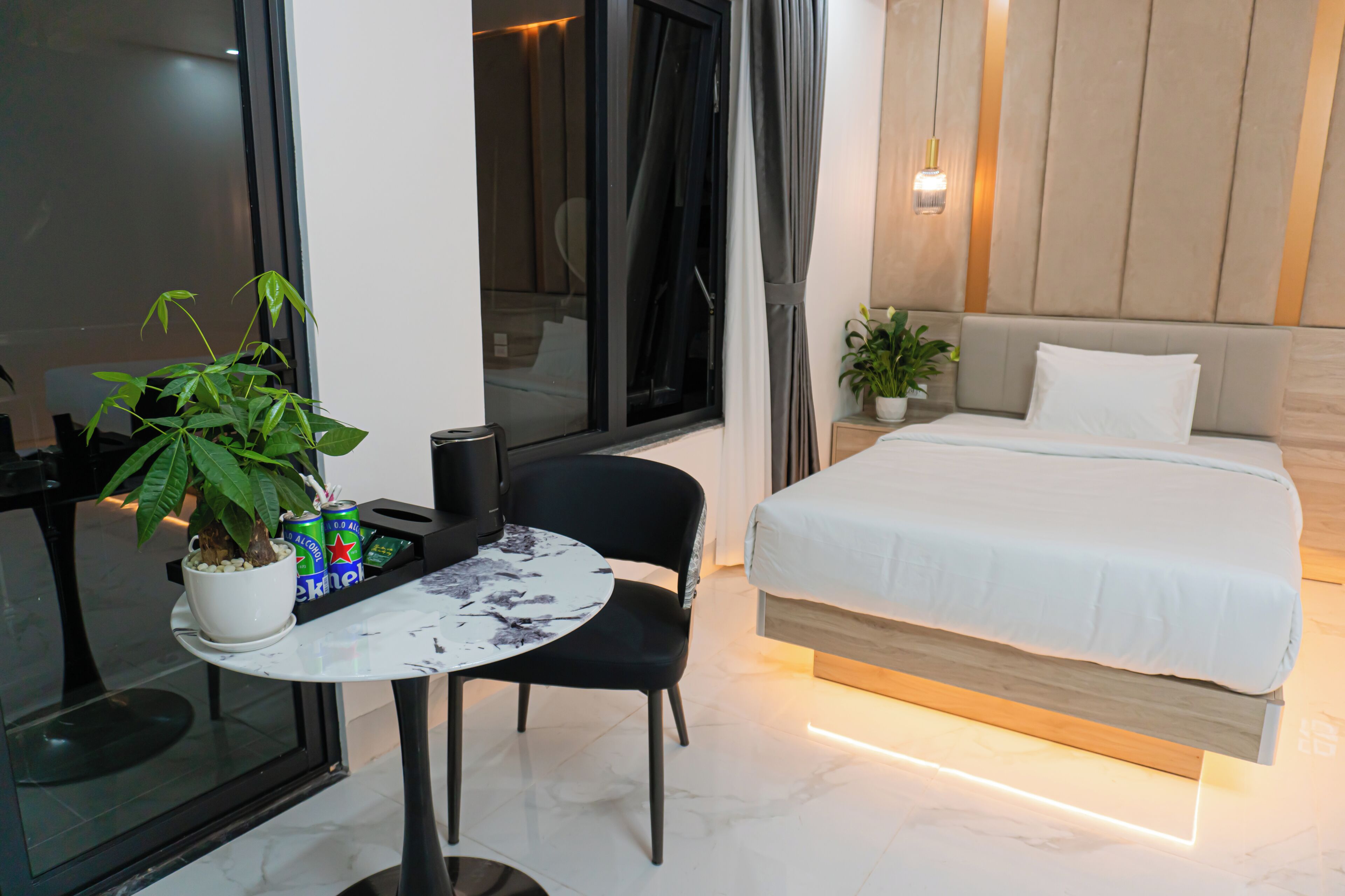 A hotel room with a bed, a small table with a potted plant, a chair, and a window with curtains.