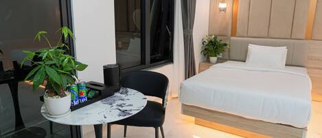 A hotel room with a bed, a small table with a potted plant, a chair, and a window with curtains.