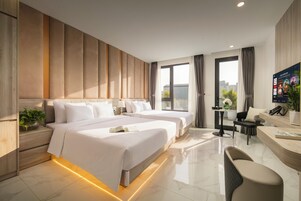 Quarto de hotel com duas camas, uma escrivaninha, uma cadeira e uma janela grande com cortinas.