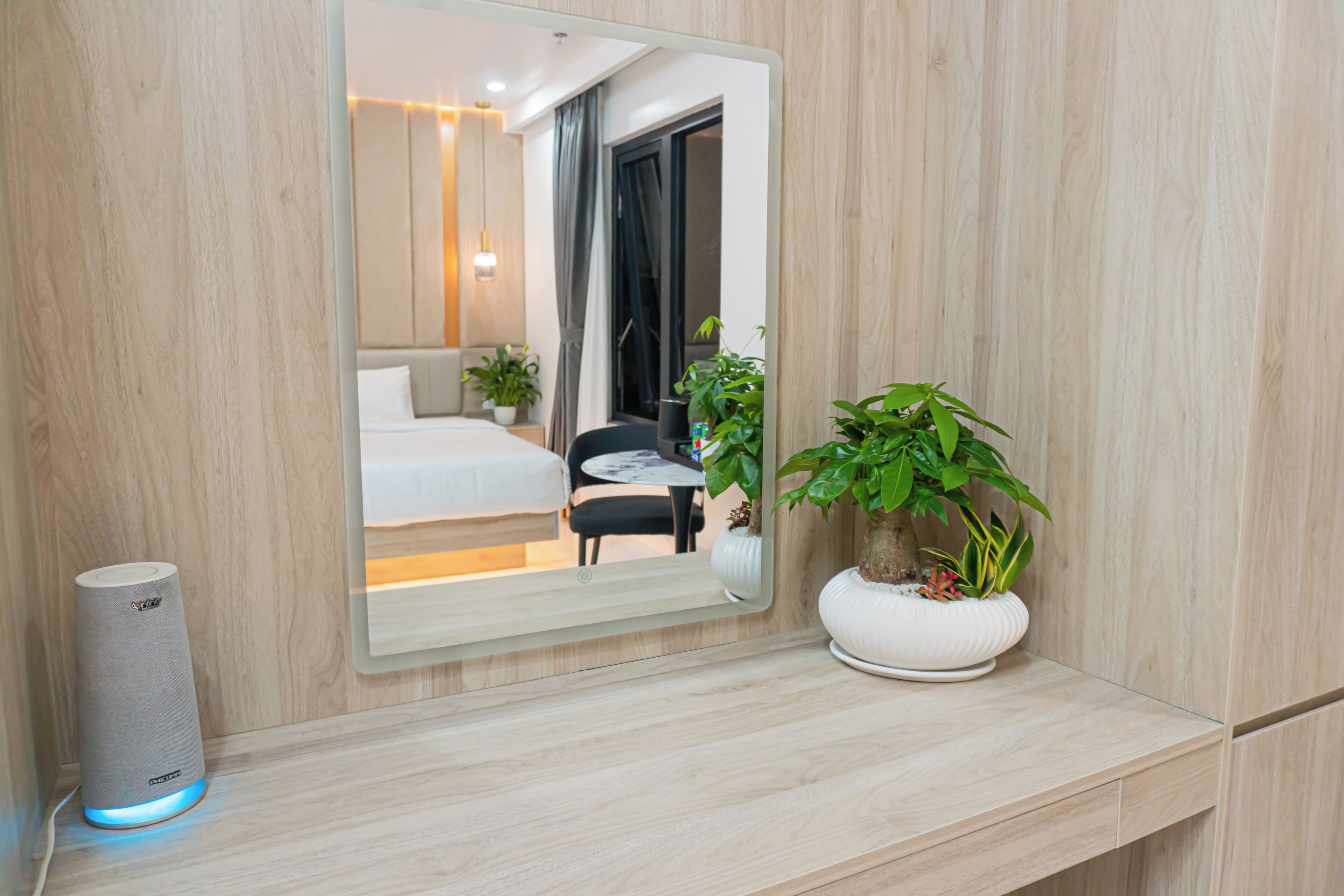 A modern hotel room with a large mirror, a bedside table with a potted plant, a bedside speaker, and a view of the bedroom through the mirror.