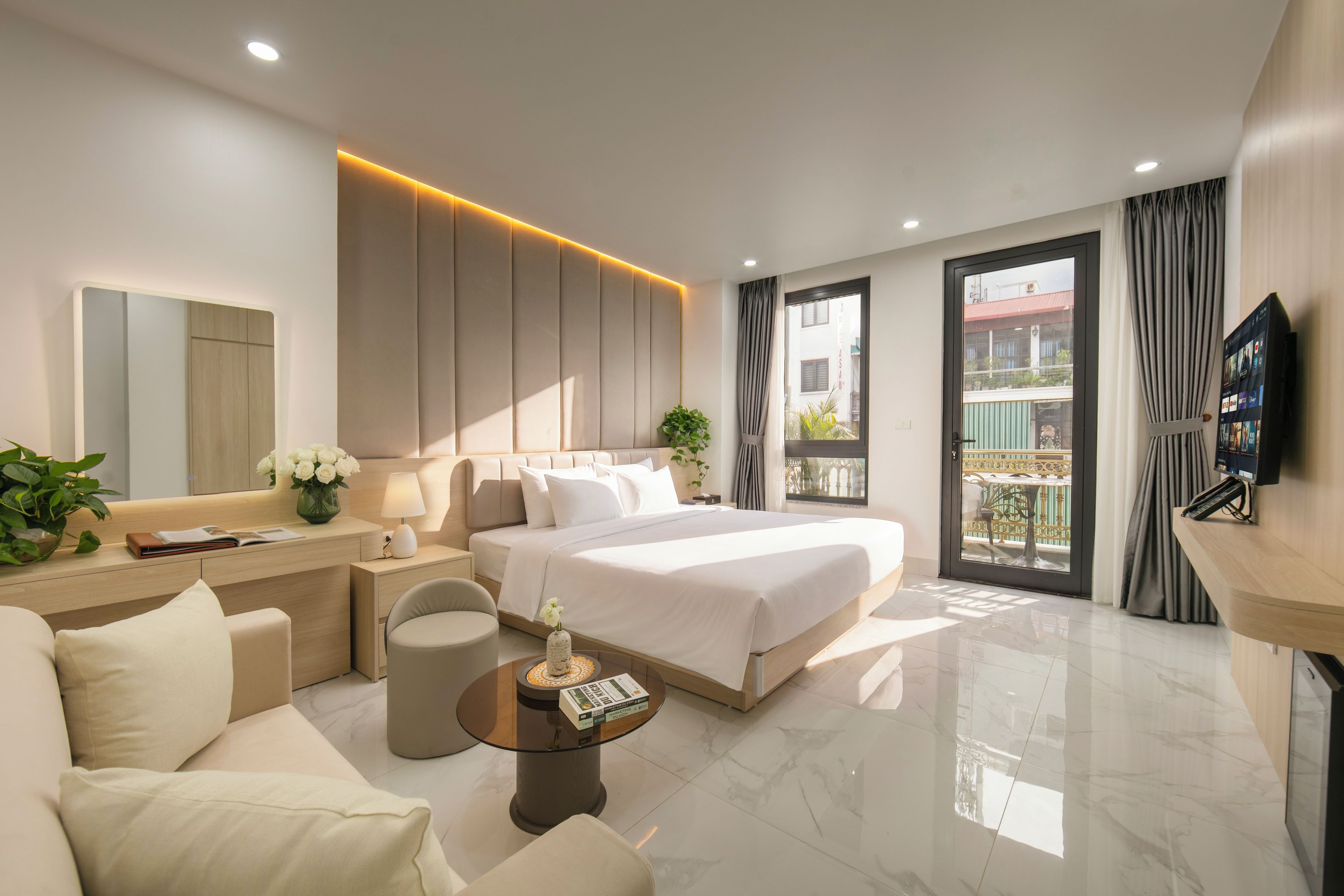 A modern hotel room with a bed, sofa, bedside tables, a TV, and a balcony with curtains.