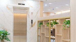 A modern lobby with a marble wall, an elevator, wooden shelves with plants, and a seating area.