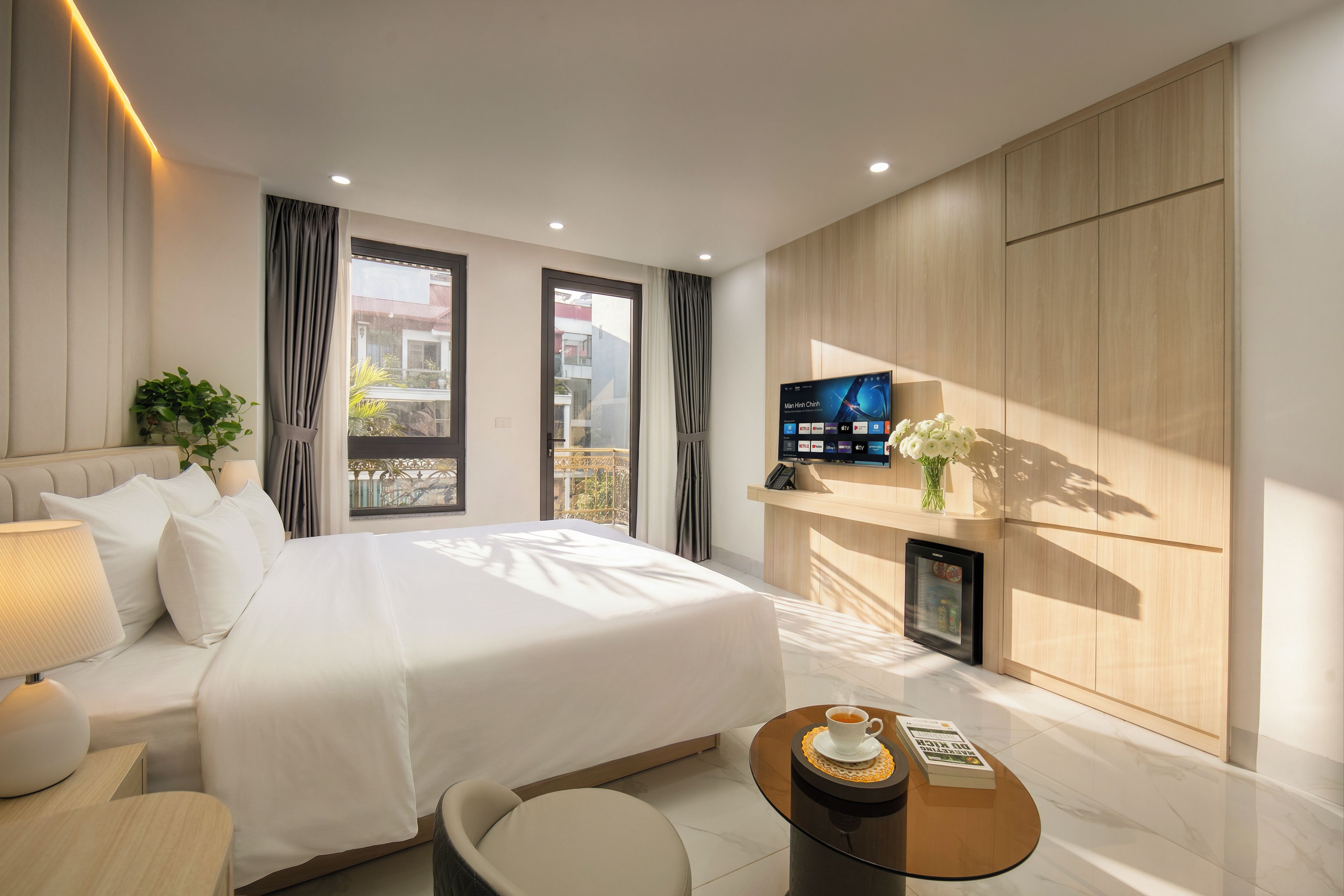 A modern hotel room with a large bed, a flat-screen TV, a small refrigerator, and a coffee table.