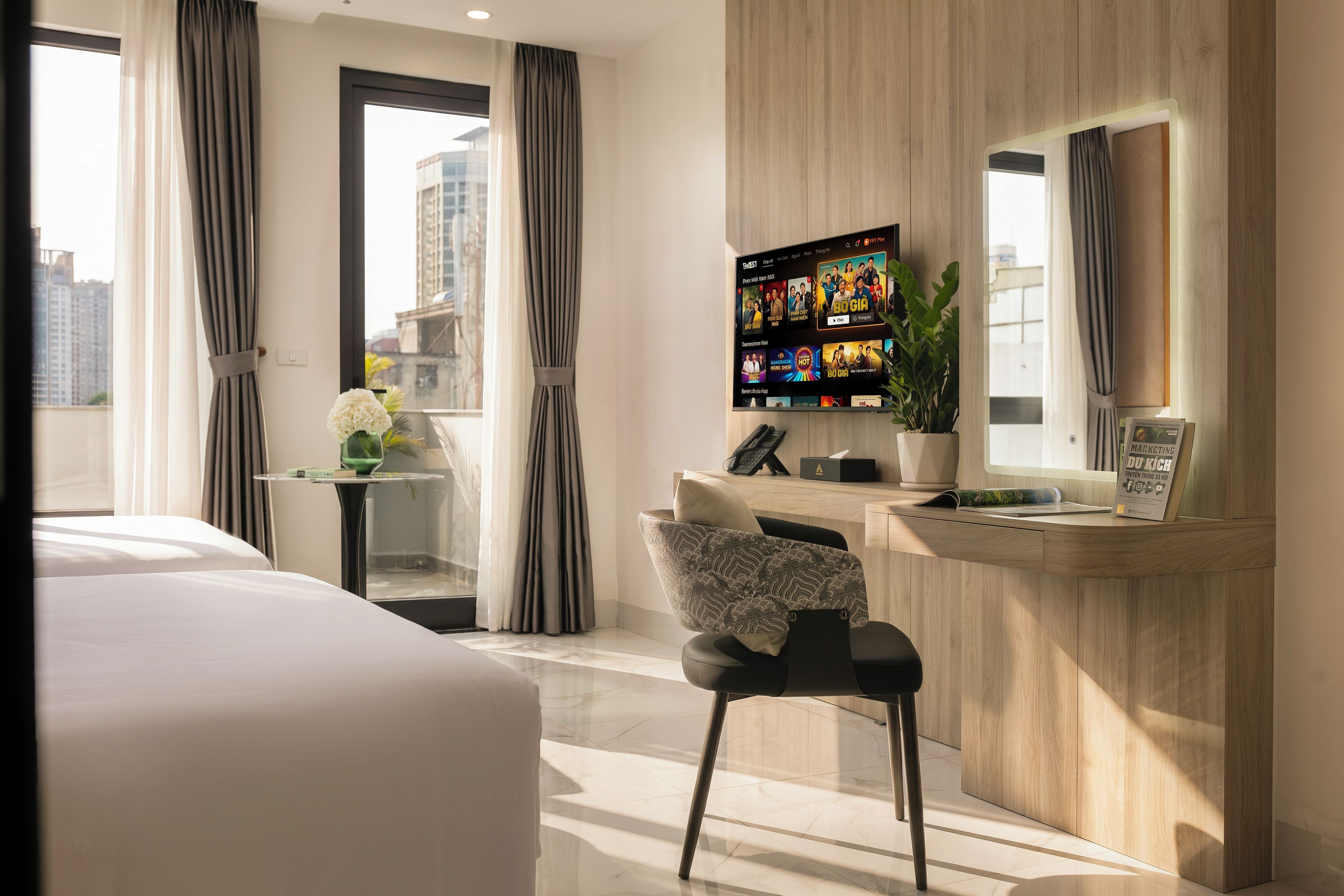 A hotel room with a bed, a desk with a computer, a chair, a potted plant, and a TV displaying a movie.