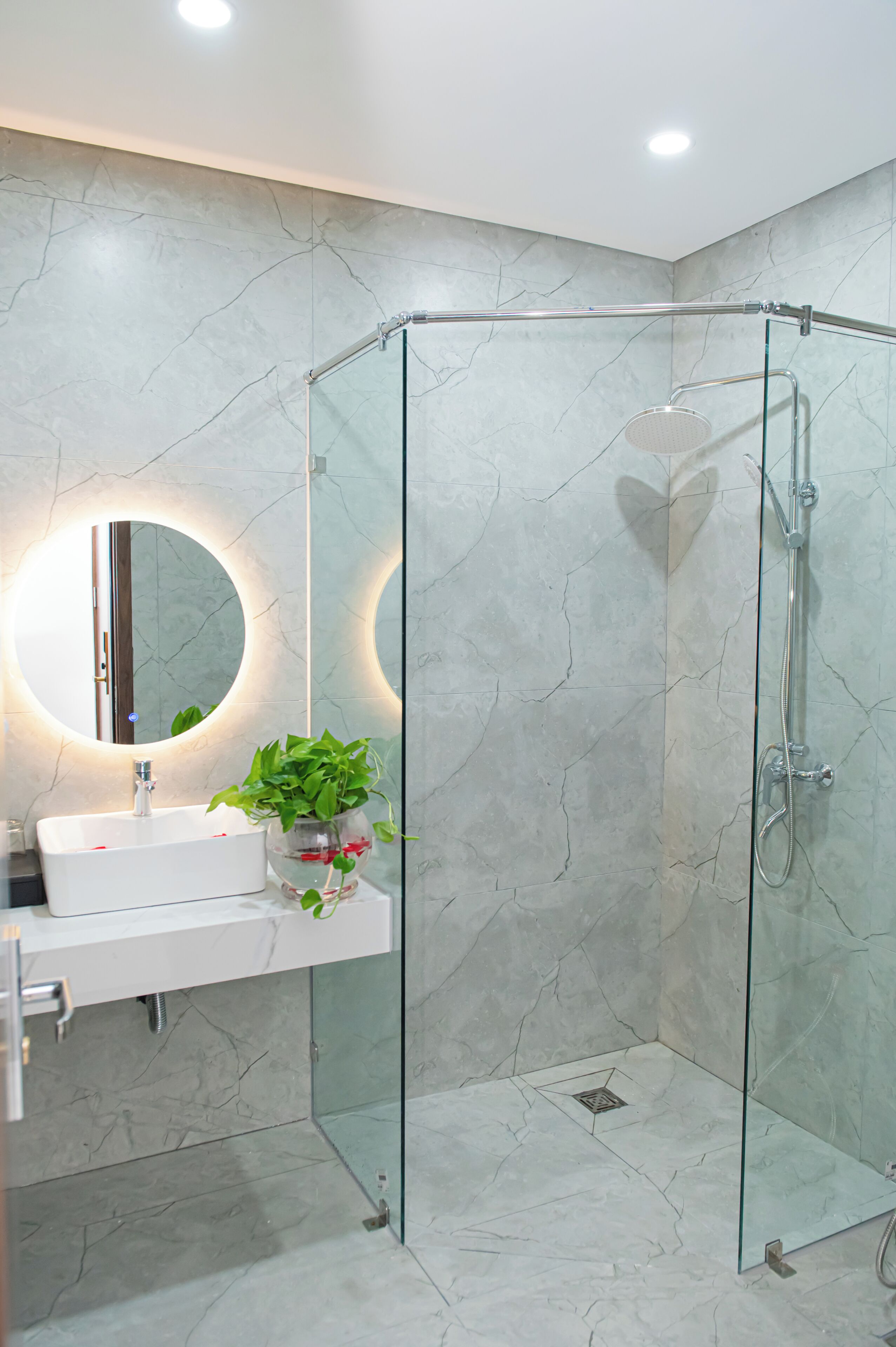 A modern bathroom with a glass-enclosed shower, a round mirror, a white sink, and a potted plant.