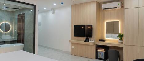 A modern hotel room with a bed, a desk with a flat-screen TV, a bathroom with a mirror and sink, and an air conditioning unit.
