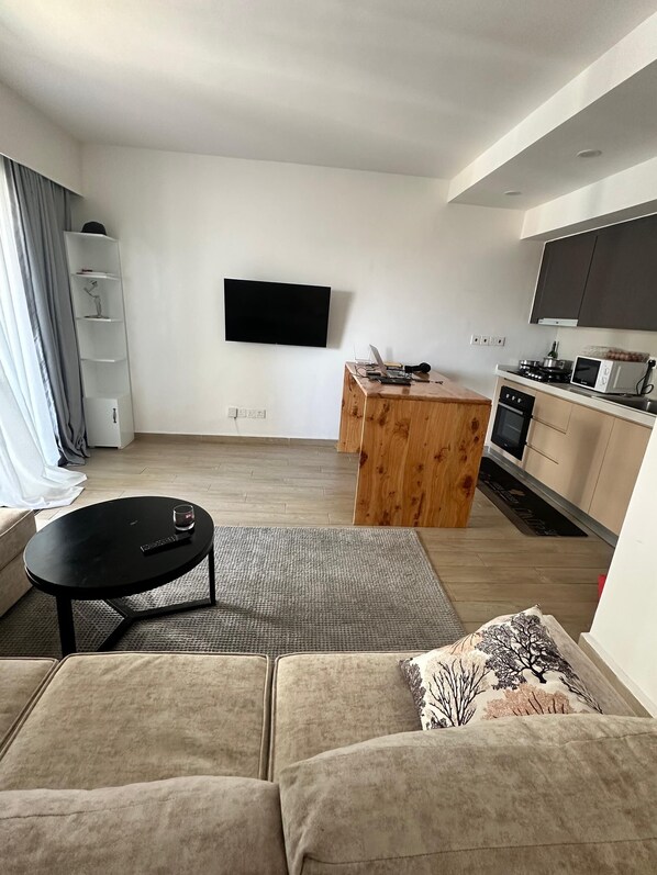 A modern living room with a sofa, a coffee table, a TV mounted on the wall, a wooden dining table, and a kitchen area with a microwave and stove.