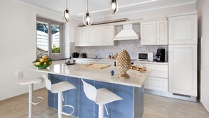 Cozinha moderna com armários brancos, uma ilha azul com banquetas, iluminação pendente e vista para o exterior.
