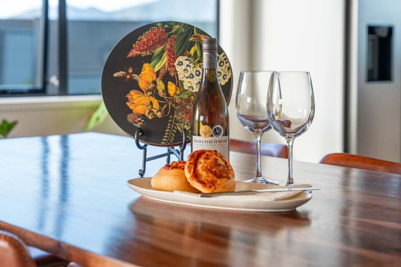 A dining table with a bottle of Old Coach Road wine, two glasses, a plate with food, and a decorative plate with a floral design.