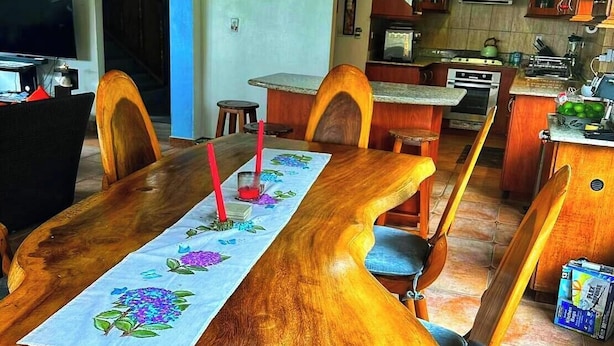 Une table à manger recouverte d’une nappe à motifs floraux, des chaises en bois et, au fond, une cuisine.