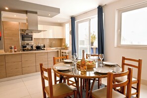A modern kitchen with built-in appliances, a dining area with a round table and chairs, and a large window with curtains. - Modern & Terrace by the Beach, 3brd in Qawra (St. Paul's Bay)